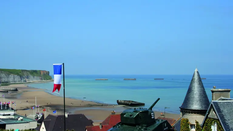 A coastal town above a wide sandy beach with concrete Mulberry-harbour blocks in the sea, a tank and tricolour in the foreground.