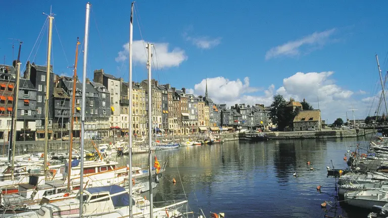 A picturesque harbour of tall slate-fronted houses reflected in calm water with sailing boats.