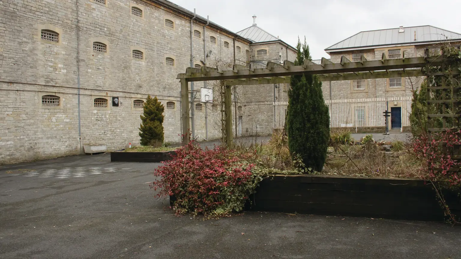 Tall stone wing of a Victorian prison with rows of small barred windows, beside an overgrown courtyard and a wooden pergola