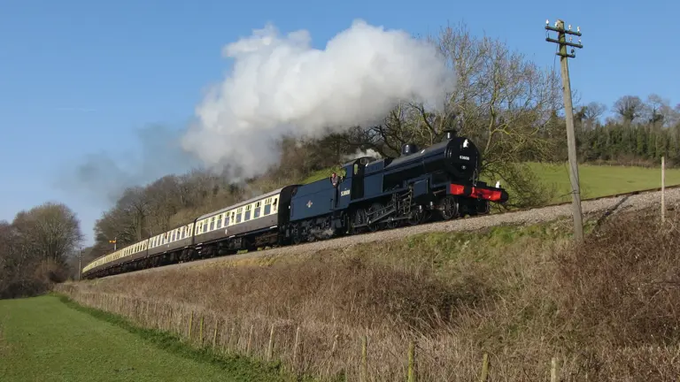 Black steam locomotive hauling a long line of cream and brown carriages along a country embankment beneath a clear blue sky