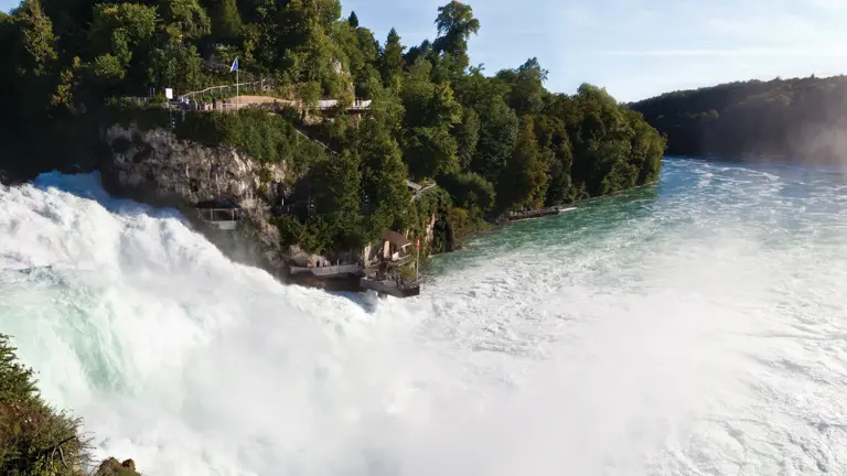 The wide Rhine Falls thundering over a rocky ledge into a turquoise river, a small castle perched on the wooded cliff above