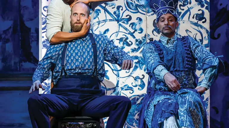 Figaro shaving a wide-eyed customer in a barber's chair, watched by a figure in an elaborate plumed headdress, against blue and white tiled scenery, in Glyndebourne's production of The Barber of Seville