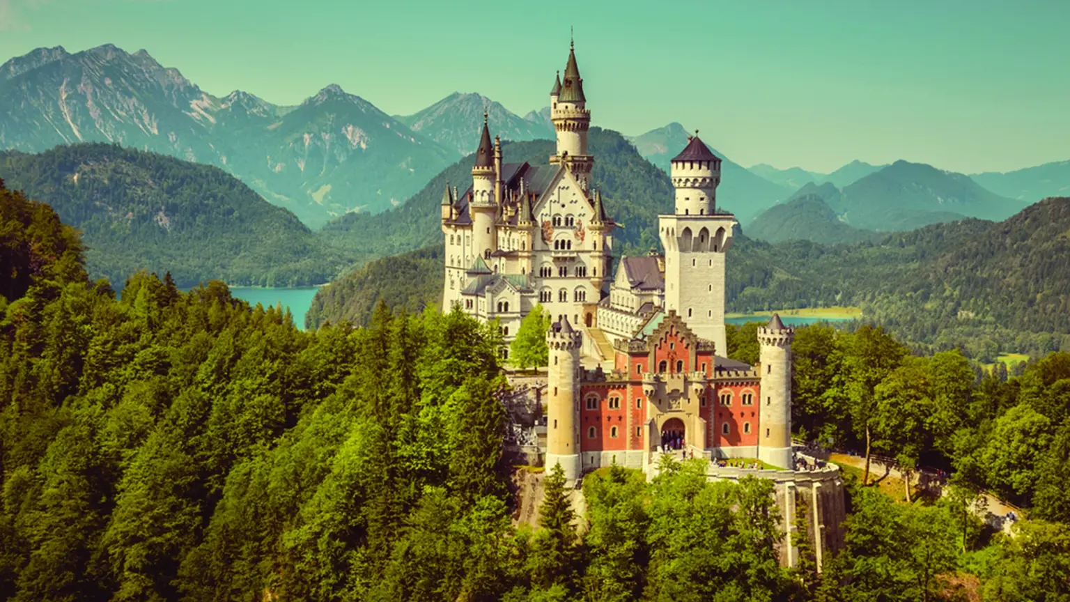 The white turreted Neuschwanstein Castle perched on a forested crag with the Bavarian Alps rising behind