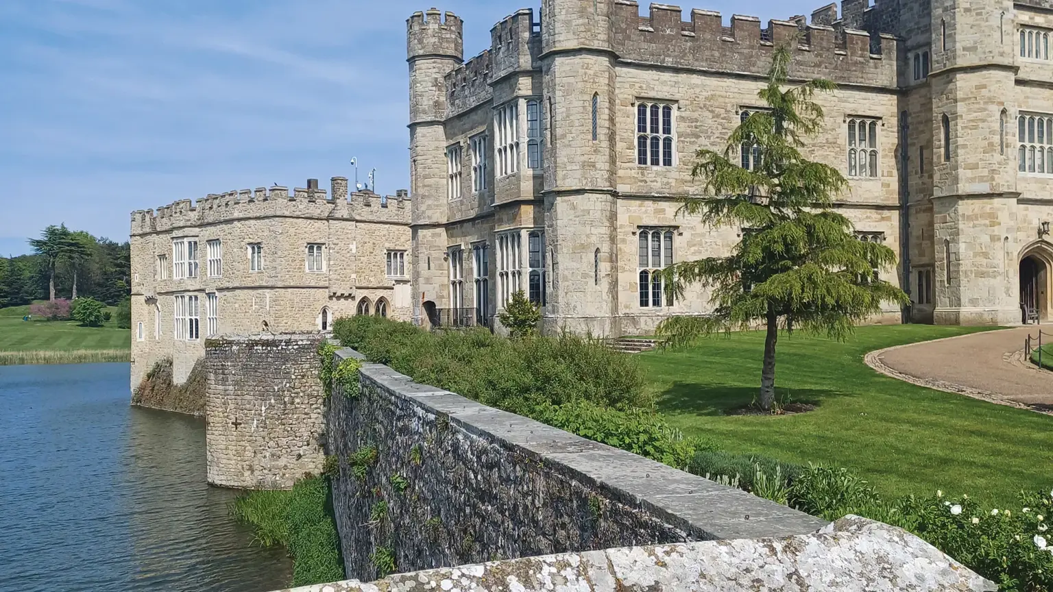Pale stone castle with round towers rising directly from a wide moat, set in green parkland under a blue sky