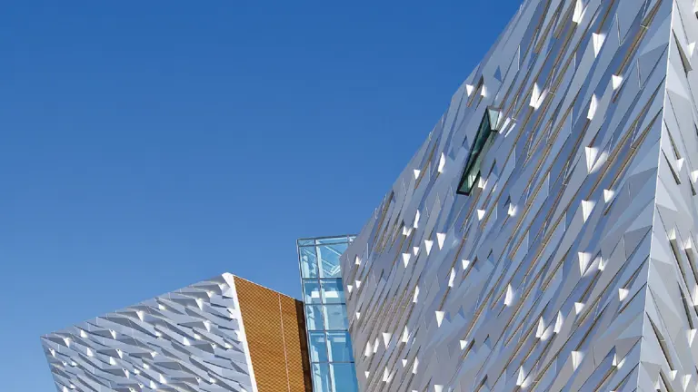 The angular silver hull-shaped facade of the Titanic Belfast building against a deep blue sky, the word TITANIC in steel letters at its base