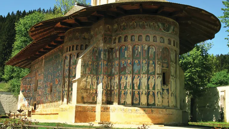 A Bucovina painted monastery church, its rounded apse and tower covered in faded blue and ochre exterior frescoes, set in a garden