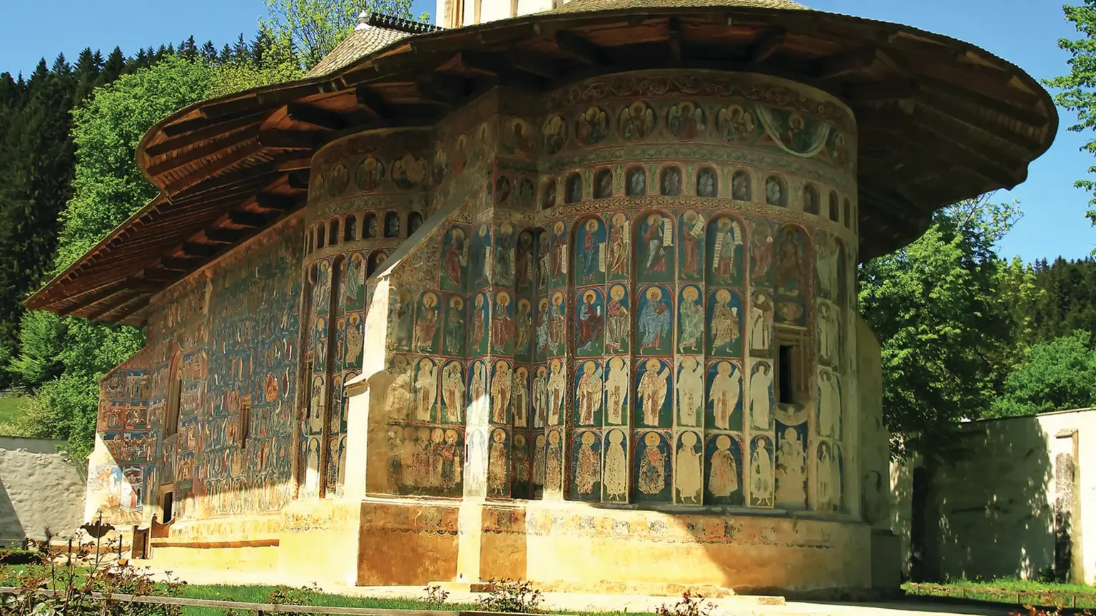 A Bucovina painted monastery church, its rounded apse and tower covered in faded blue and ochre exterior frescoes, set in a garden