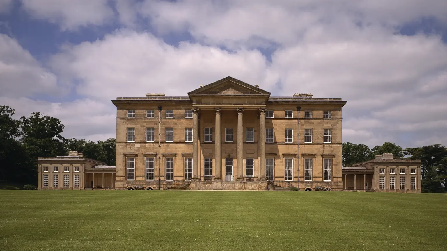 South facade of Attingham Park, a classical 18th-century mansion with pedimented portico and tall Ionic columns, set behind a sweeping lawn under a cloudy sky