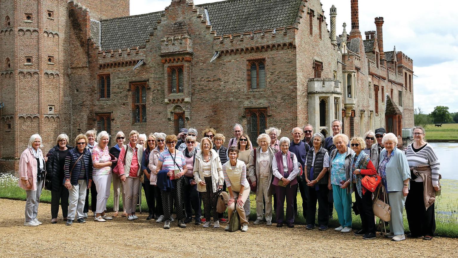 Group of visitors standing outside a large brick manor house with moated grounds on a garden visit.