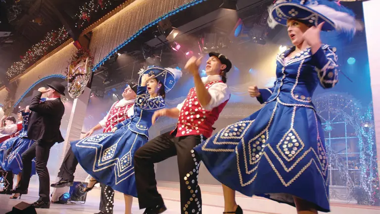 Dancers in blue and red sequinned costumes and feathered hats performing under stage lights
