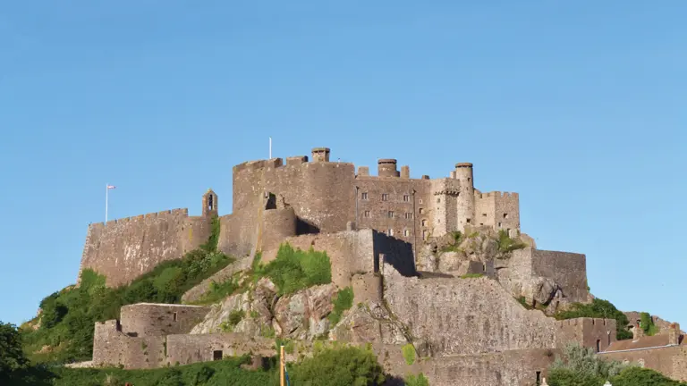 The medieval Mount Orgueil Castle on its rocky hilltop above Gorey harbour, with boats grounded on the low-tide sand and seafront houses below