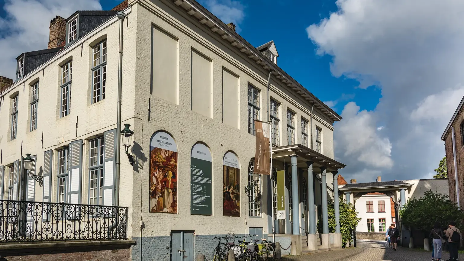 A pale neoclassical museum building with a small pillared porch and painting banners on its facade, bicycles parked outside.