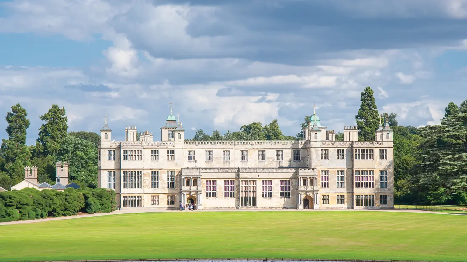 Grand Jacobean country house in pale stone with symmetrical wings, multiple chimneys and a central courtyard, set behind sweeping lawns
