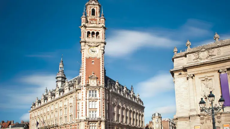 A tall Flemish-Renaissance belfry with a clock beside a classical opera house on a paved square under blue sky.