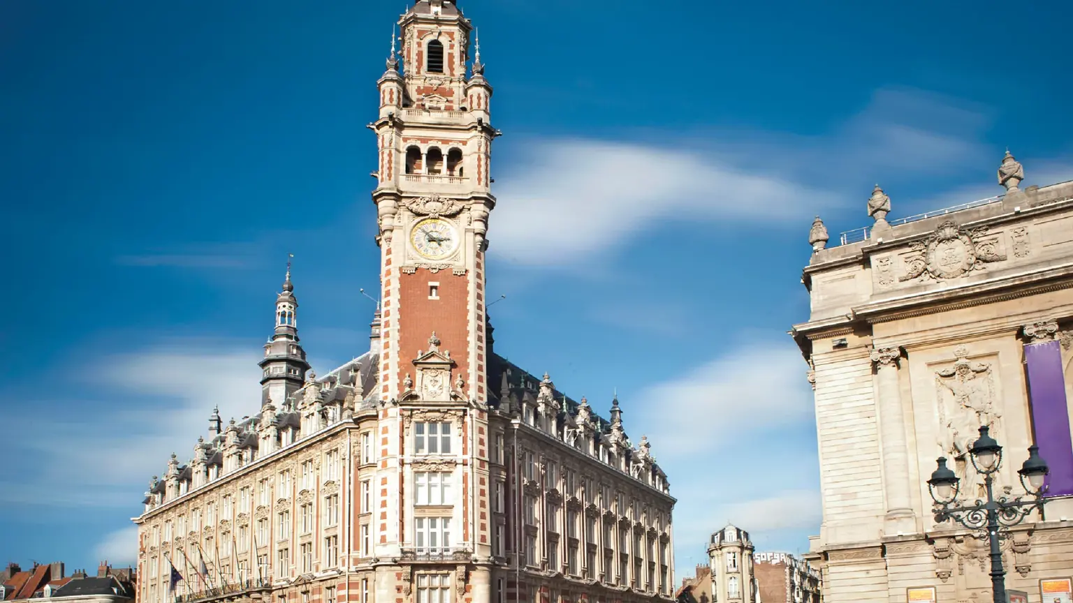 A tall Flemish-Renaissance belfry with a clock beside a classical opera house on a paved square under blue sky.