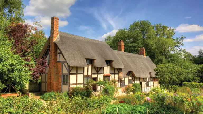 Picturesque thatched Tudor cottage with whitewashed walls and timber framing, surrounded by traditional English flower garden