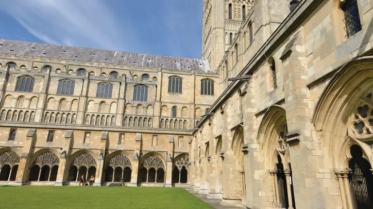 Norman cathedral with a tall slender stone spire, viewed across a cloister garth with cropped lawn