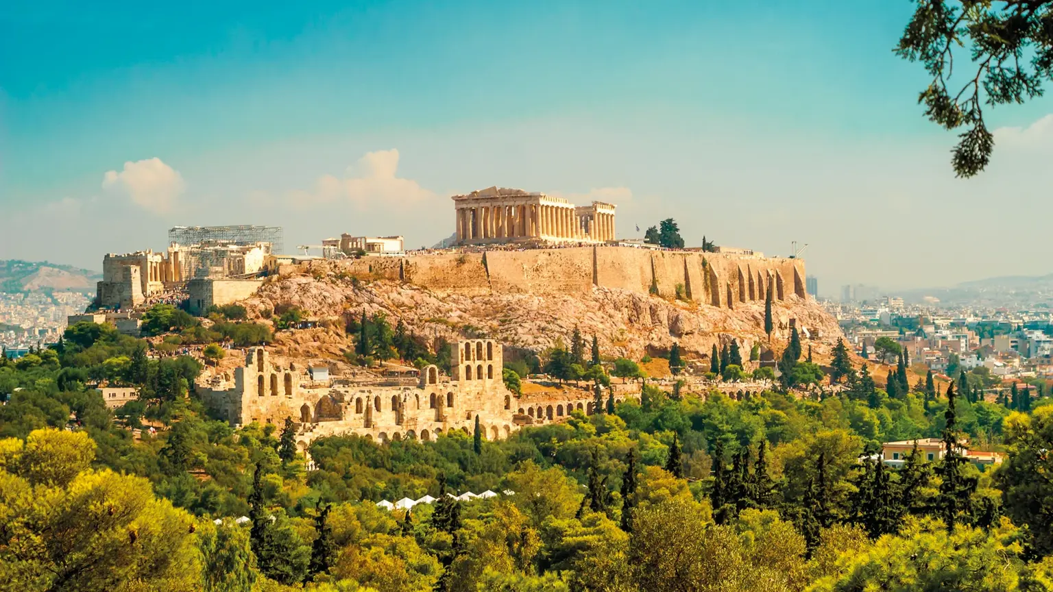The Acropolis of Athens rising above the city, with the Parthenon on the summit and the Odeon of Herodes Atticus on the slope below, framed by green trees