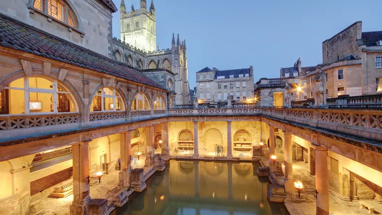 The torchlit colonnaded Great Bath of Bath at dusk, with the tower of Bath Abbey rising behind