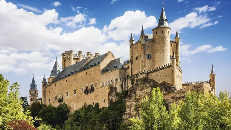 The Alcázar of Segovia, a fairytale castle with pointed slate turrets rising on a rocky outcrop above wooded slopes