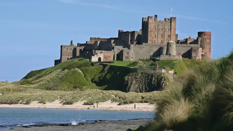 Bamburgh Castle on a rocky promontory above a sweeping sandy beach on the Northumberland coast, set against a clear blue sky