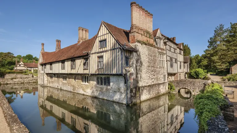Timber-framed medieval manor house with red-tiled roofs reflected in a still moat, surrounded by trees