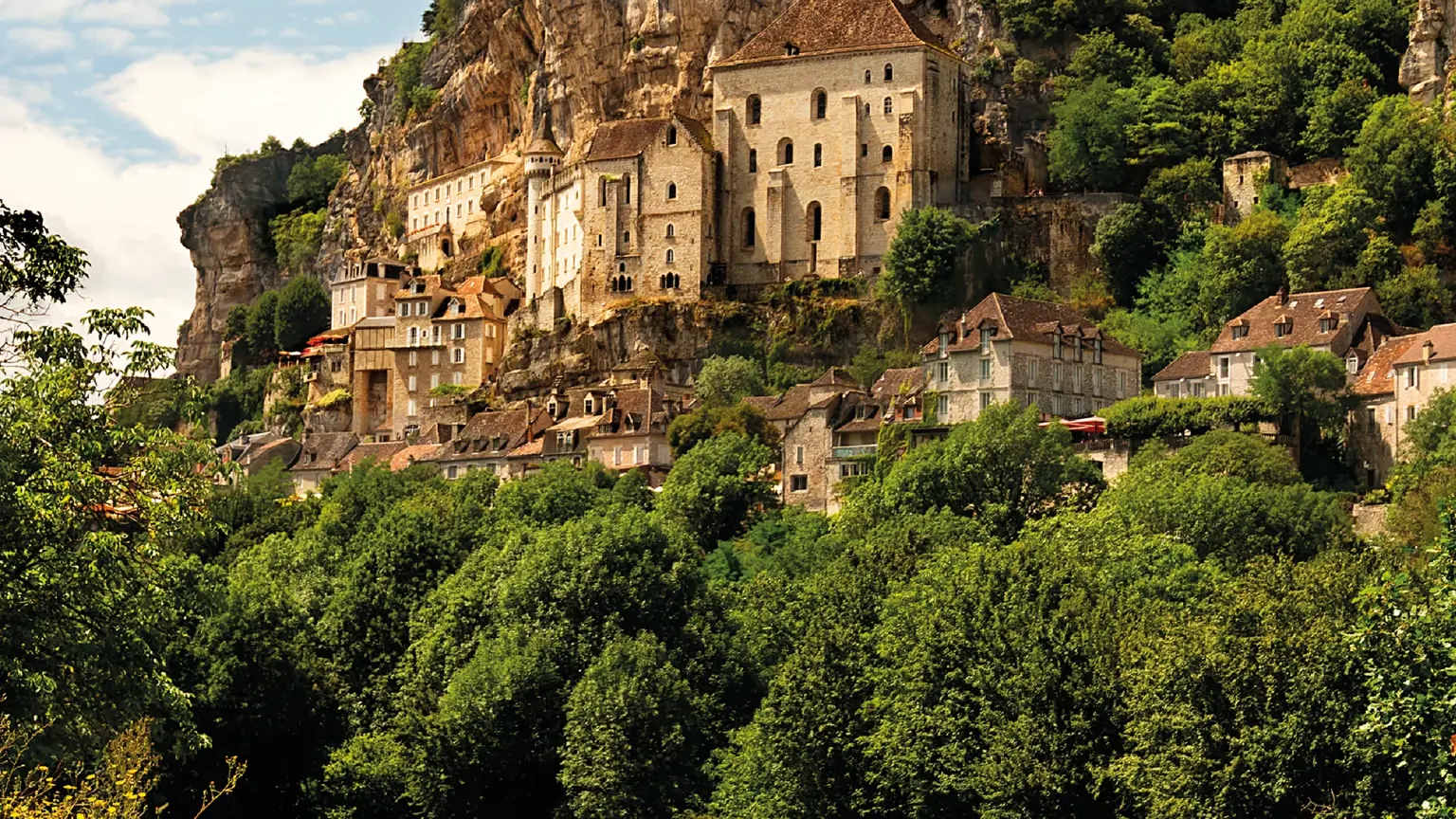 Houses, chapels and a castle stacked vertically up a tall limestone cliff above a green meadow.