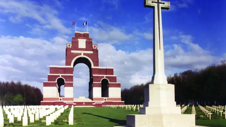 A massive red-brick and stone arch memorial beside a tall white cross and rows of war graves.