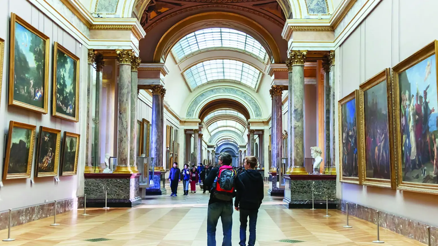 A long arched gallery with skylights, marble columns and walls of gilt-framed paintings, visitors walking through.