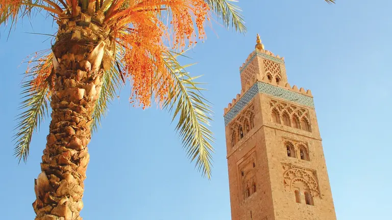 The minaret of the Koutoubia Mosque in Marrakech rising against a clear blue sky, framed by the fronds of a date palm in the foreground