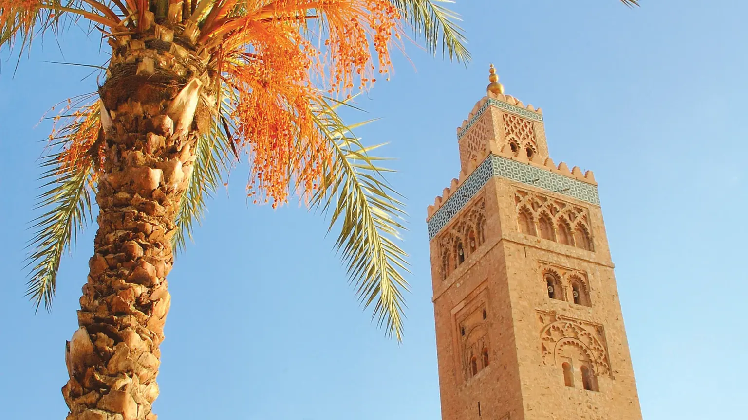 The minaret of the Koutoubia Mosque in Marrakech rising against a clear blue sky, framed by the fronds of a date palm in the foreground