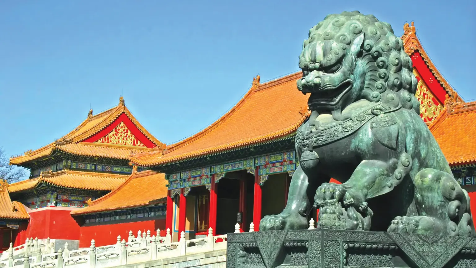 A bronze guardian lion in front of the red walls and golden tiled roofs of the Forbidden City in Beijing