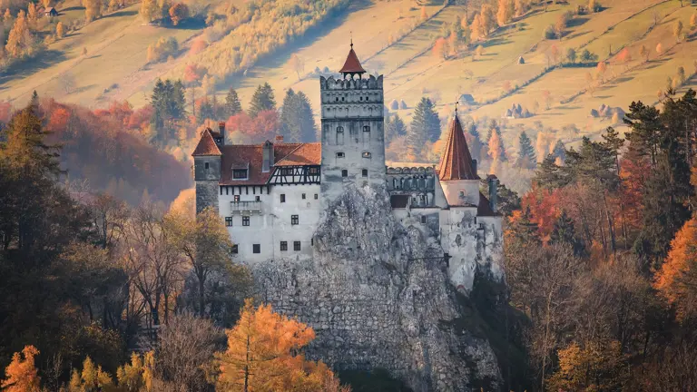 Bran Castle on its rocky bluff, a white-walled fortress of towers and red roofs against forested mountains, popularly known as Dracula's Castle