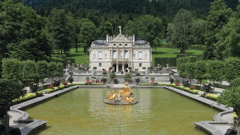 Linderhof Palace beyond its formal garden pool and golden fountain statue, wooded Alpine slopes rising behind