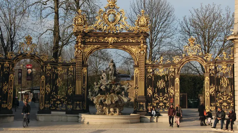 Black-and-gold ornate iron gates flanking a sculpted fountain on a cobbled square.
