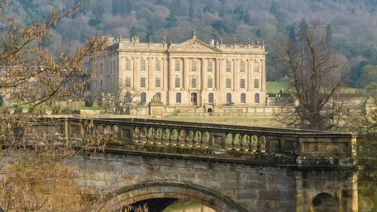 Honey-stone Palladian Chatsworth House framed by trees, viewed across a graceful arched stone bridge over the River Derwent
