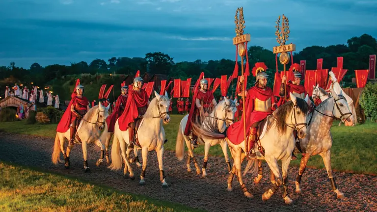 Riders in Roman costume with red cloaks and SPQR standards ride grey horses at dusk during the Kynren outdoor show