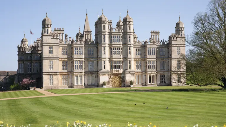 The west front of Burghley House on a bright day, the grand Elizabethan stone facade and skyline of towers and chimneys reflected in calm water in the foreground.