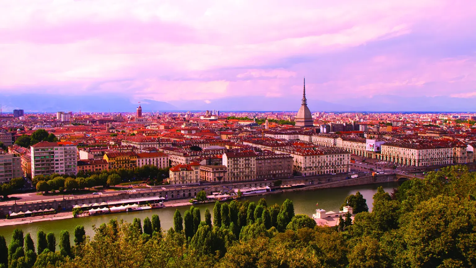 Aerial sunset view of Turin spread along the Po river, terracotta roofs and church spires with the Mole Antonelliana's distinctive aluminium spire and the Alps in the distance