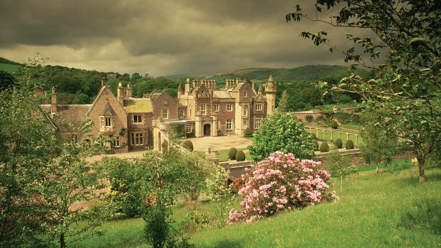The romantic Scots Baronial silhouette of Sir Walter Scott's Abbotsford House on the banks of the River Tweed, set among orchards and gardens under a brooding Borders sky
