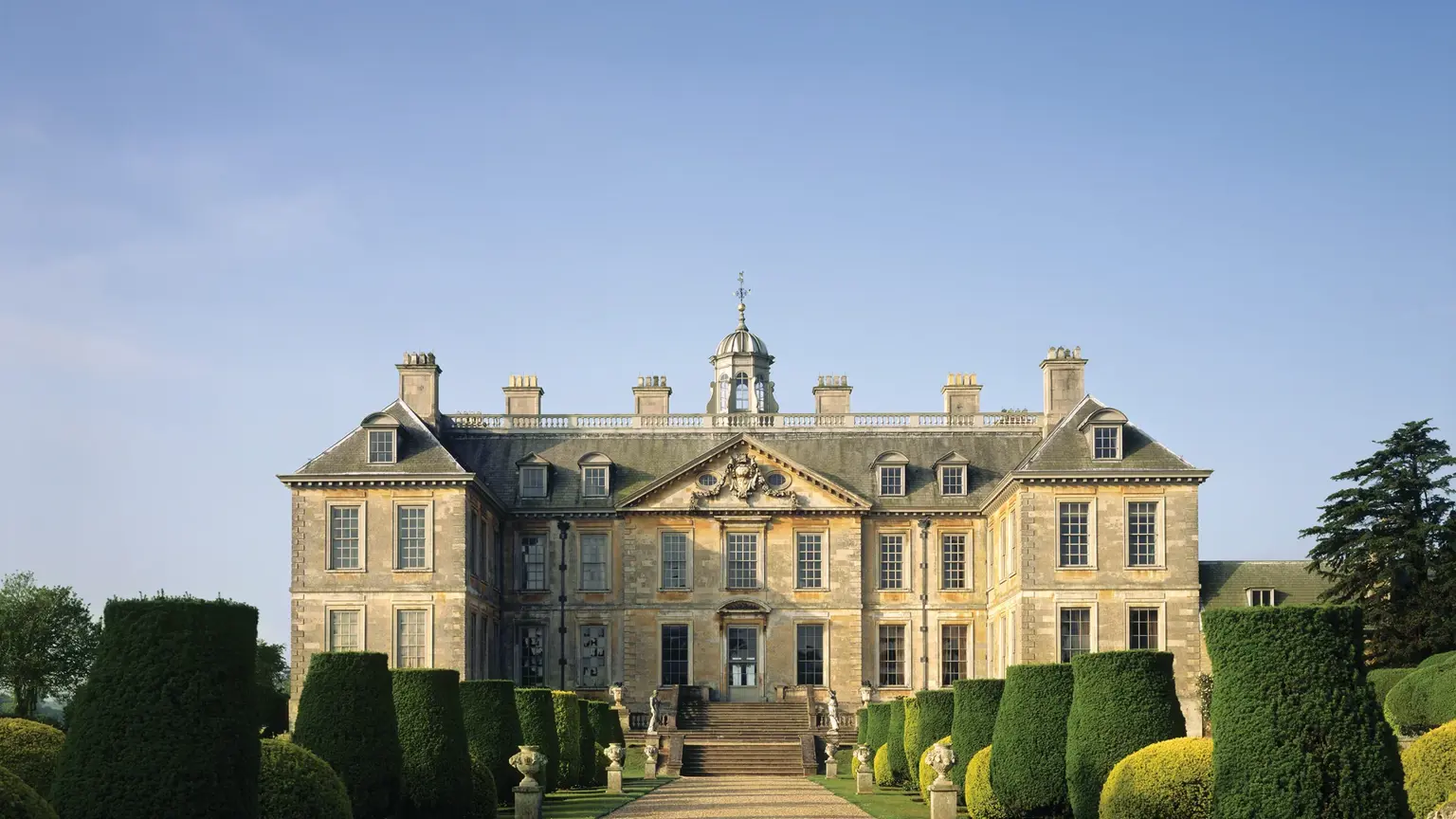 Late seventeenth century country house in honey-coloured stone, symmetrical wings either side of a central pediment, set behind a wide manicured lawn
