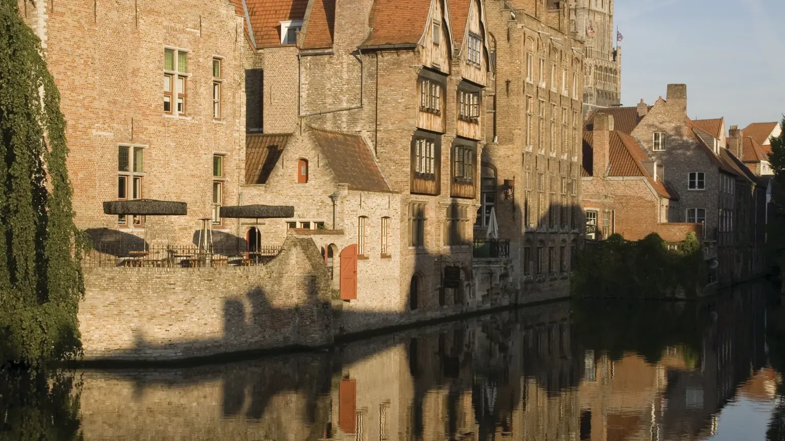 Stone and brick canal-side houses at a bend of water with a tall medieval belfry behind, all mirrored in the still canal.