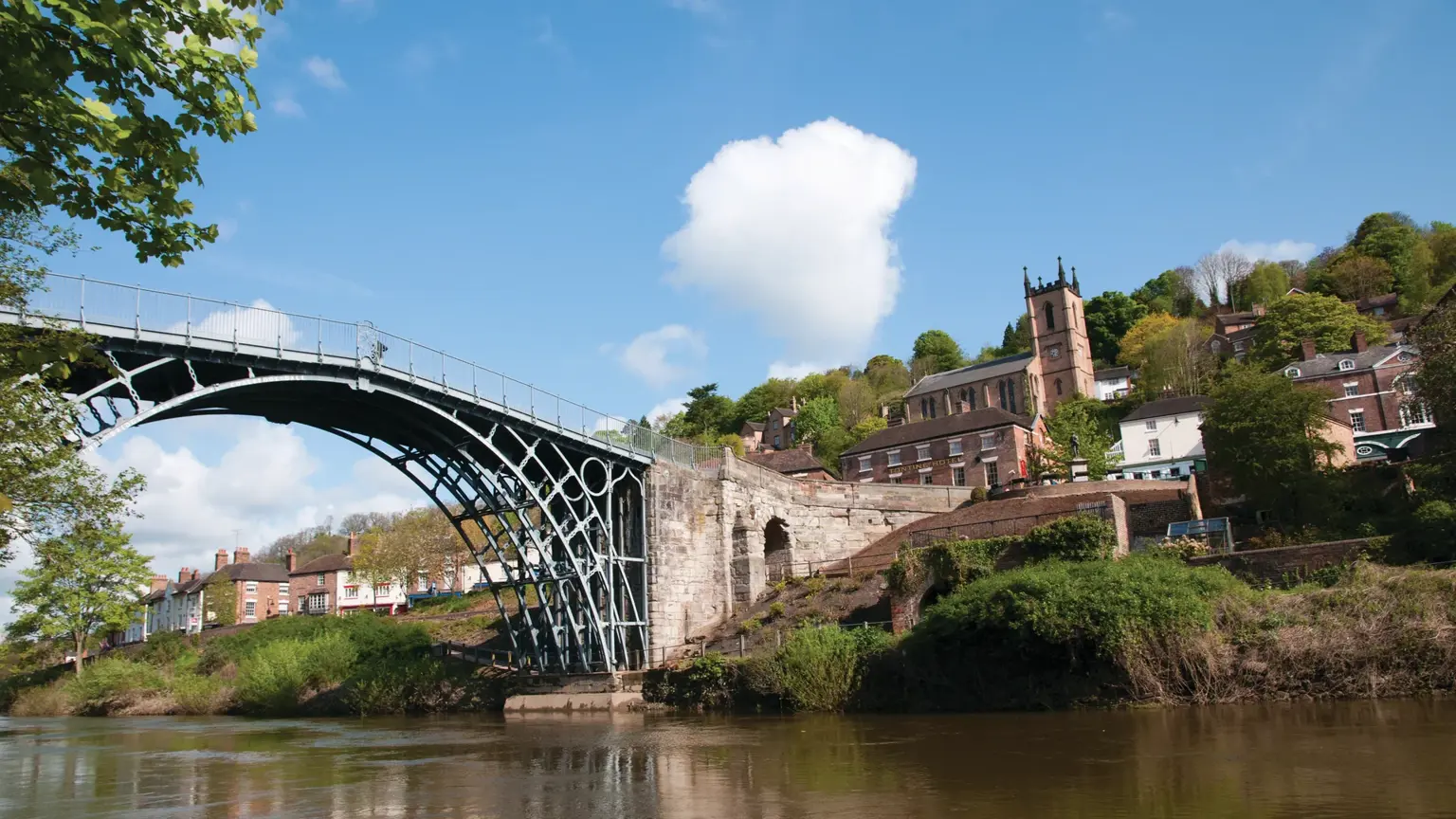 Graceful single-span cast-iron bridge arching over the River Severn, with the riverside village of Ironbridge rising up the wooded gorge behind