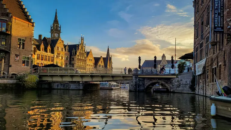 Medieval cityscape with historic stone buildings and bridges reflected in calm waterfront, blue sky with clouds above