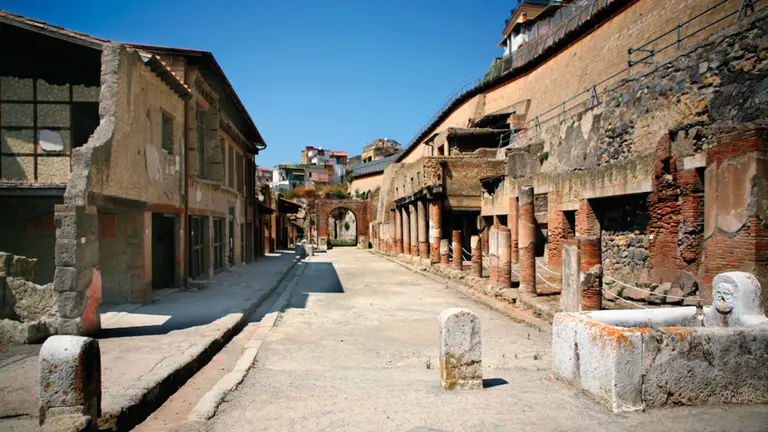 The excavated streets of Herculaneum, brick-and-stone Roman buildings exposed below the modern city above, the Bay of Naples beyond