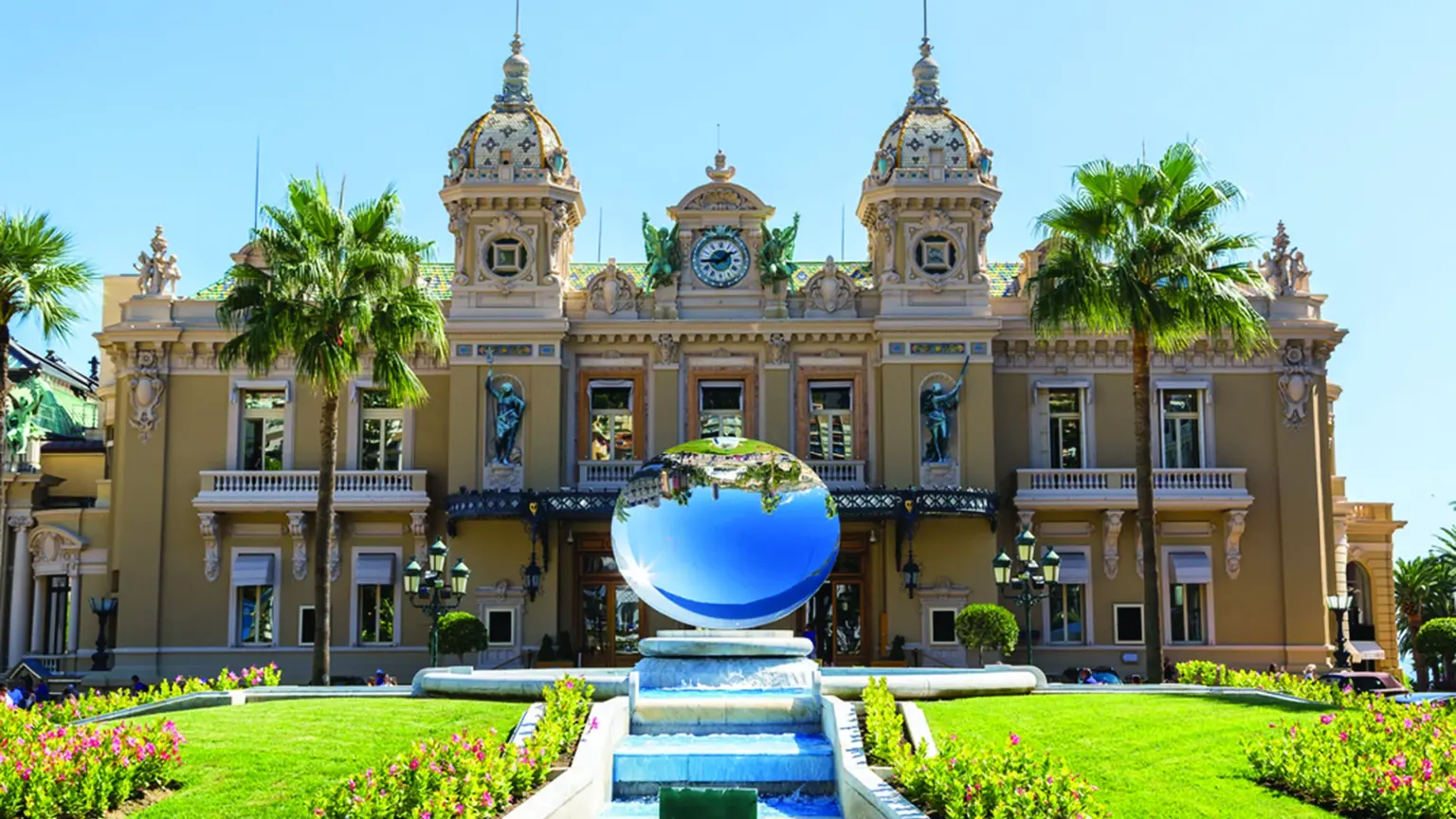 The Beaux-Arts facade of the Monte Carlo Casino in Monaco, twin ornamental domes above the formal gardens and a blue mirror-sphere sculpture in a reflecting pool