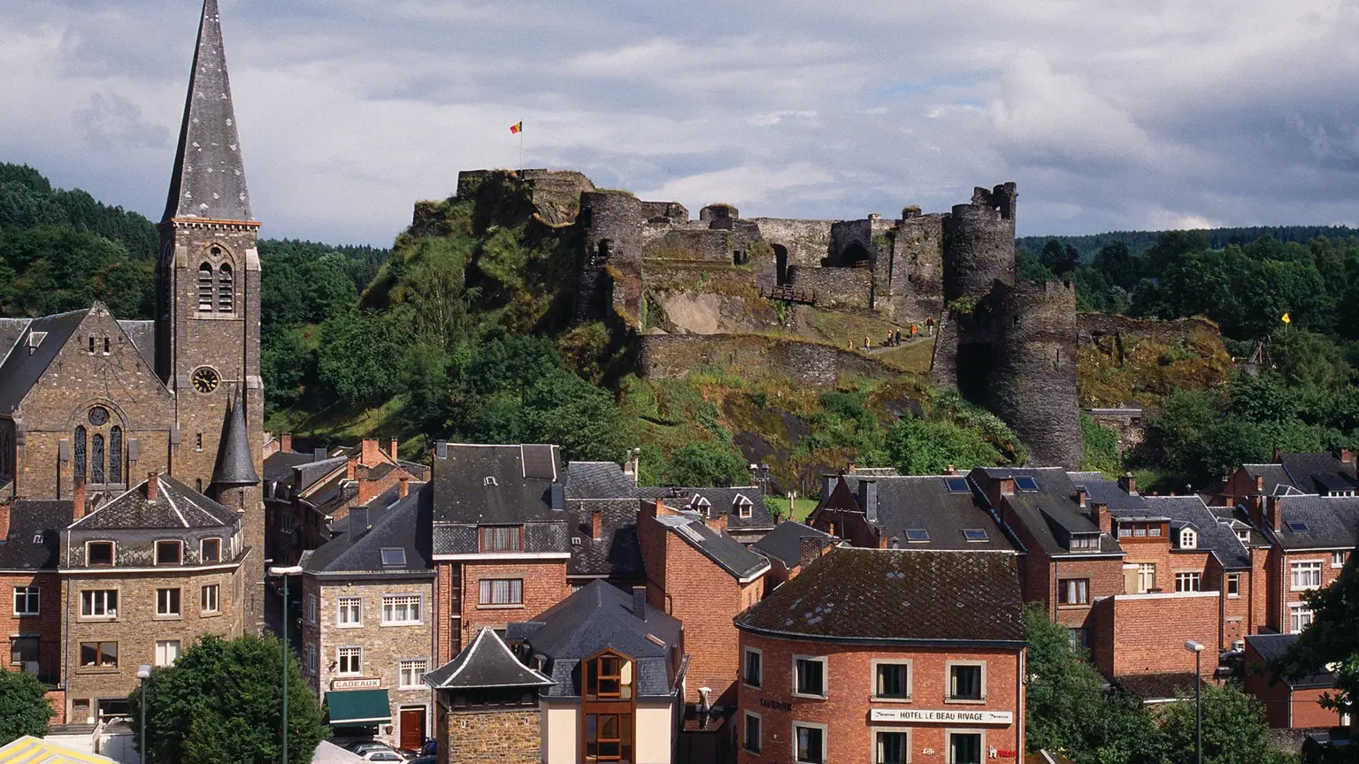 A small Ardennes town of slate-roofed houses and a church spire below a craggy hilltop crowned by ruined stone castle walls flying a flag.