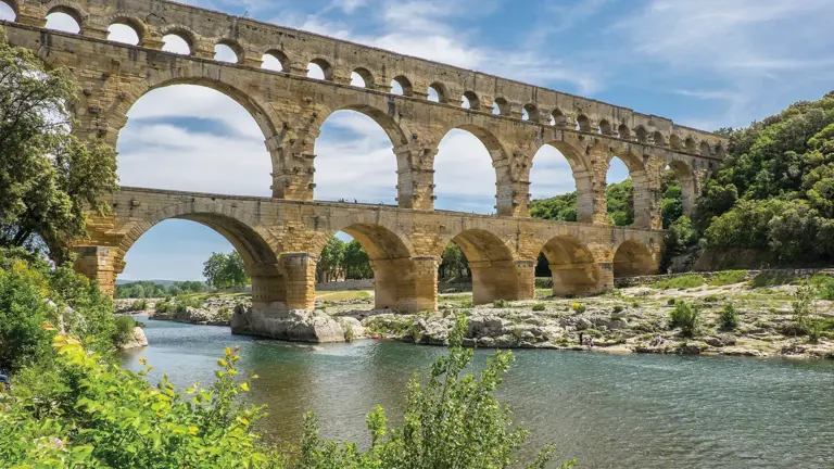 A honey-coloured three-storey Roman arched aqueduct spanning a green river under blue sky.