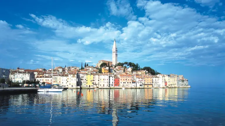 The colourful waterfront of Rovinj rising to the bell tower of St Euphemia's church, its pastel houses reflected in the calm harbour water