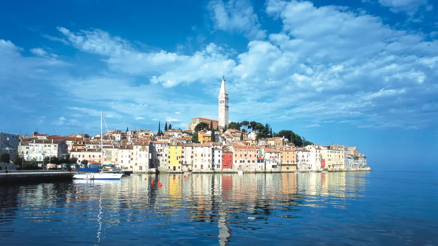 The colourful waterfront of Rovinj rising to the bell tower of St Euphemia's church, its pastel houses reflected in the calm harbour water